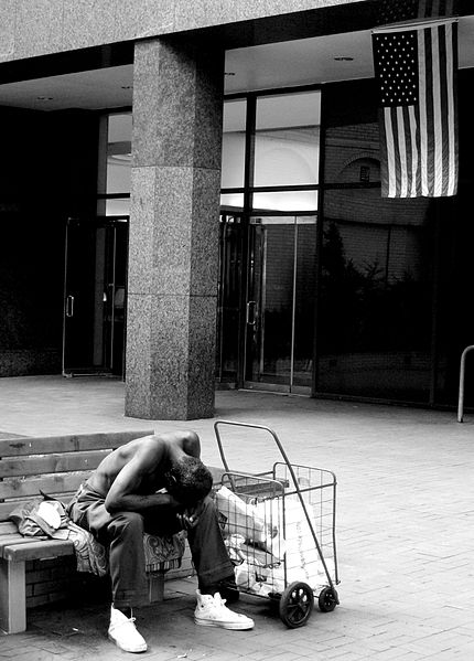 A homeless man outside the outside the United Nations building in New York with the American flag in the background. By C. G. P. Grey 